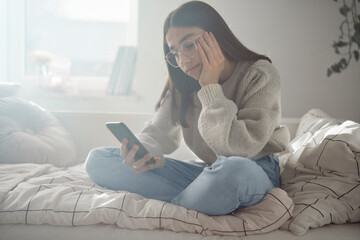 Bored caucasian teenage girl browsing phone while sitting on bed