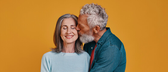 Senior man kissing hid smiling wife while standing together against colored background