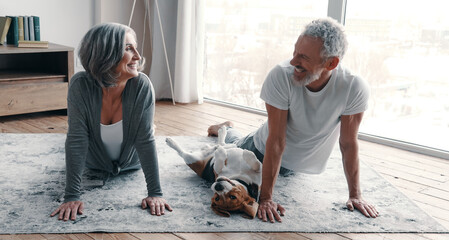 Cheerful senior couple practicing yoga with their dog at home together