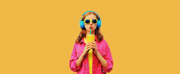 Portrait of stylish young woman listening to music in headphones with cup of fresh juice wearing pink jacket on orange background