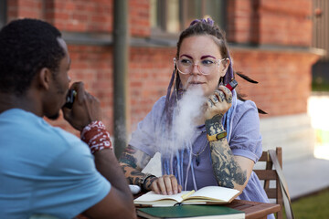 Young businesswoman looking at her colleague while both smoking vape pens and discussing working points at outdoor business meeting
