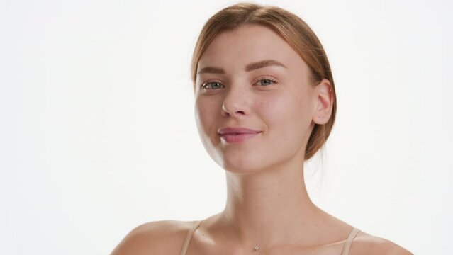 Face care cosmetics commercial | Close-up shot of cute young slim brown-haired Caucasian woman strokes her cheek looking at the camera and smiling for it on white background