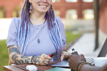 Close-up of young smiling businesswoman with vape pen networking at meeting with African American male colleague in outdoor cafe