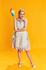 Housekeeper. Portrait of beautiful young blonde woman with stylish hairstyle posing with pipidastre against yellow studio background. Concept of retro fashion, beauty, 50s, 60s. Pin-up style