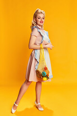 Grocery shopping. Portrait of beautiful young girl with stylish hairstyle posing with net bag against yellow studio background. Concept of retro fashion, beauty, 50s, 60s. Pin-up style