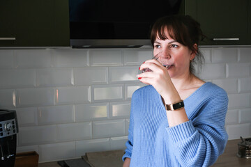 A young woman is drinking clean water from a glass tumbler in her kitchen.