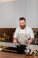 Bearded man smiling and cooking in pot on modern stove in kitchen.