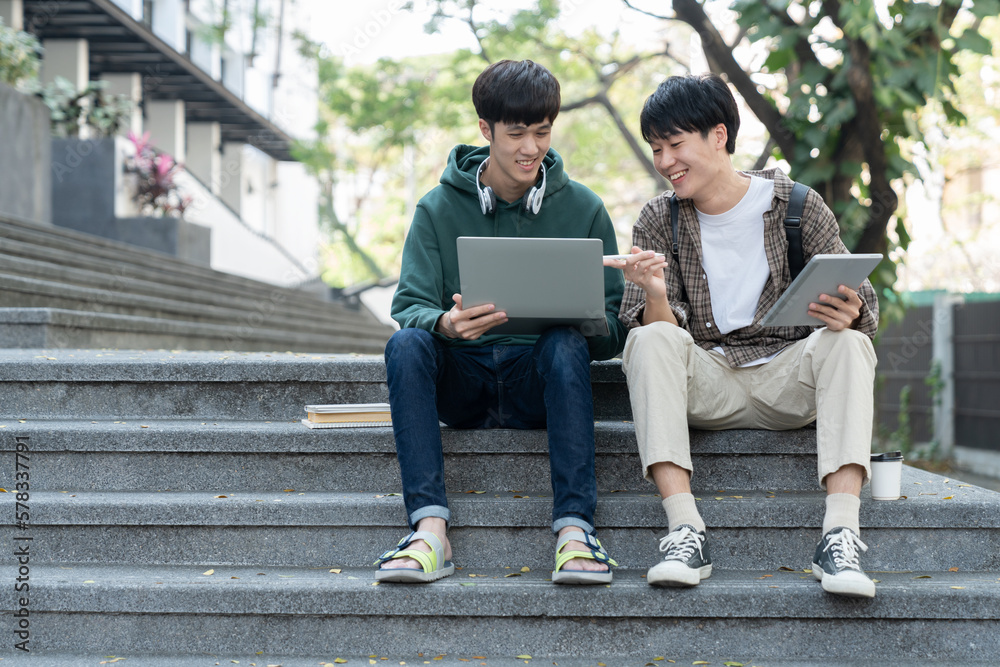 Wall mural two asian male students sitting on the stairs of the university enjoy chatting after school using sm