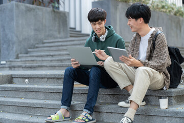Two Asian male students sitting on the stairs of the university enjoy chatting after school using smartphones and tablets to find information together.