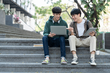 Two Asian male students sitting on the stairs of the university enjoy chatting after school using...