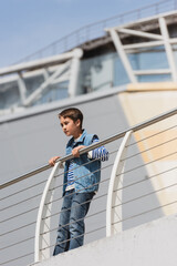 low angle view of stylish boy in denim vest and jeans standing near metallic fence on embankment.