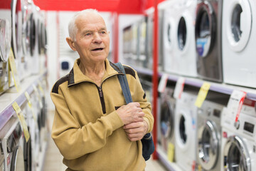 elderly grayhaired man pensioner looking washing machine at counter in showroom of electrical...