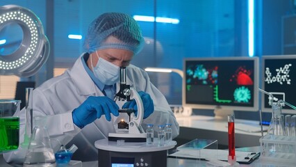 A male researcher in a white gown, mask, blue gloves and a bonnet is examining a sample in a microscope. People in biochemical overalls is working in the background.