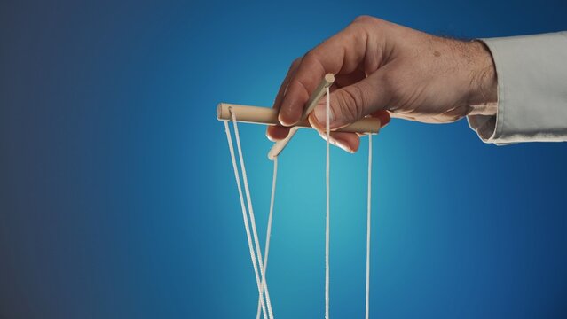 The hand of a man in a gray shirt controls a puppet using a wooden manipulator and strings on a blue background. Hand handling at puppet by pulling strings to make the character move. Close up.