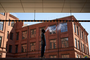 Side view of smoking young black man in casualwear standing against red brick building and...