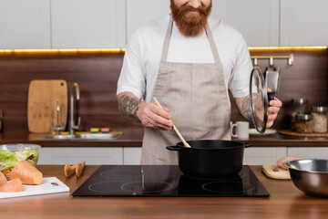 Cropped view of bearded man cooking in pot near salad on worktop in kitchen.