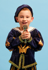 Happy preschool age boy in costume of medieval pageboy, little prince singing at microphone over...