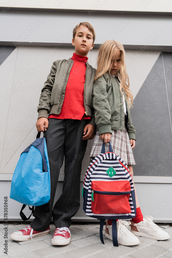 Poster low angle view of stylish kids in bomber jackets holding backpacks while standing near mall.