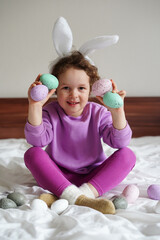 Happy Easter. cute beautiful girl with curly hair and bunny ears sitting on the bed with colorful easter eggs. Lifestyle photo