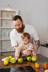 Fototapeta premium Tattooed dad holding baby bottle near daughter and fruits in kitchen.
