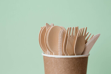 Eco-friendly disposable kitchen utensils in a paper cup on a beige-green background. Wooden forks, spoons and knives. Ecology, the concept of zero waste.
