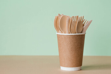 Eco-friendly disposable kitchen utensils in a paper cup on a beige-green background. Wooden forks, spoons and knives. Ecology, the concept of zero waste.