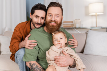 Smiling gay man hugging partner with baby daughter at home. - obrazy, fototapety, plakaty