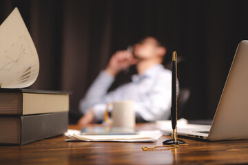 Closeup of a pen attached to a chain in stand lying on desk near laptop and books with documents with a business man sitting in blurry background in modern office cabin talking over mobile phone
