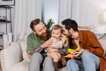 Happy gay couple holding baby girl with apple in living room.