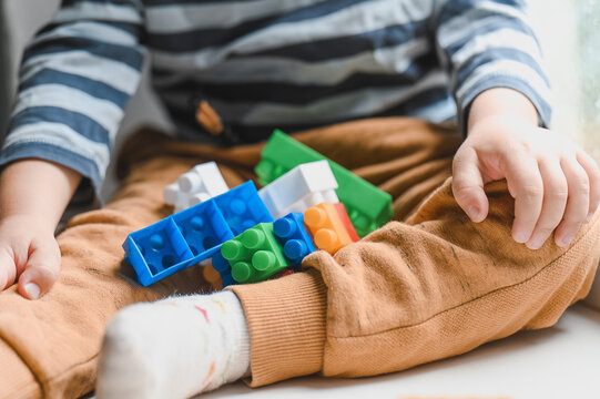 Little boy sitting on the window sill and  playing with lots of colorful plastic blocks constructor. Boy playing with construction blocks at kindergarten. No face. Close up unrecognizable