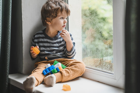 Little boy sitting on the window sill and  playing with lots of colorful plastic blocks constructor. Boy playing with construction blocks at kindergarten.  looking on the rain drops on the window
