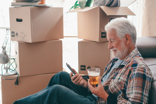 Smiling Senior Man Sitting On Floor With Glass Of Beer Using Smartphone Relaxing In New Home Living Room With Cardboard Boxes Packed With Stuff On Moving Day.