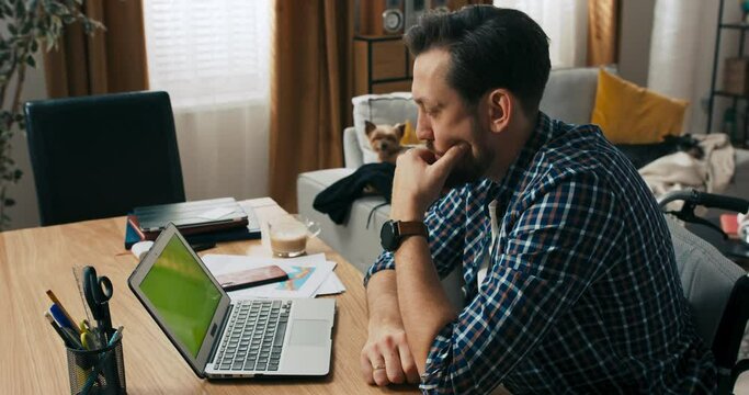 Serious concentrated young man with berd and dark hair sitting in front of coputer thinking solving problem. Man with disabilities in wheelchair working online from distance freelancer concept.