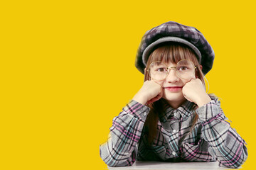 A ten-year-old girl in a plaid hat and a plaid shirt props up her chin with her hand, wearing glasses, close-up, yellow background