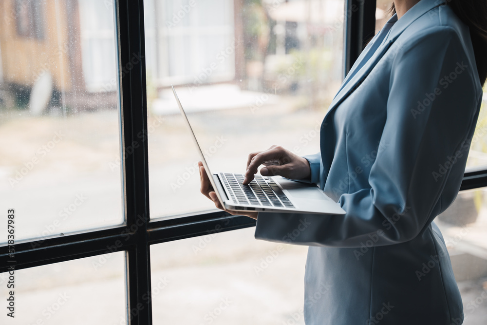 Aufkleber close up view of businesswoman holding and typing on laptop computer while standing near window.