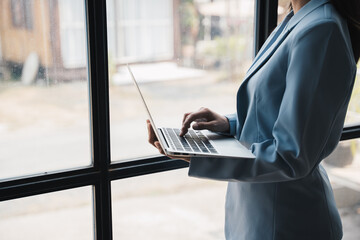 Close up view of businesswoman holding and typing on laptop computer while standing near window.