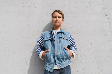 stylish preteen boy in denim vest and striped long sleeve shirt posing with hands in pockets near mall with grey wall.