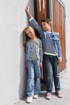 full length of stylish children in striped long sleeve shirts and denim vests posing near building.