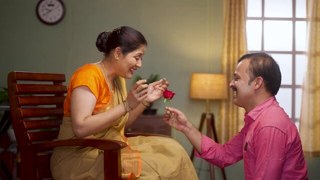 Happy middle aged man proposing his wife by giving red rose at home - concept of wedding anniversary, affection and love emotions.