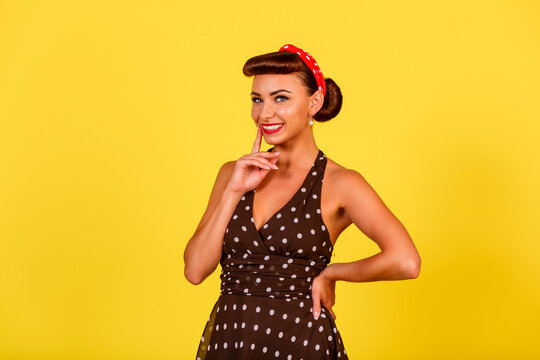 A happy smiling lady in a polka dot dress with a retro hairstyle and red lipstick stands on a yellow background. A pin-up look with a modern twist. Vintage party