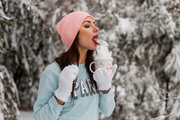 A young lady in a sweater, hat and knitted mittens in a snowy forest holds a mug of snow in her hands and licks it off. Cheerful winter mood