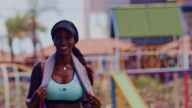 Sporty African American woman wearing sports bra, visor cap with towel around neck walking outdoors towards camera in slow motion