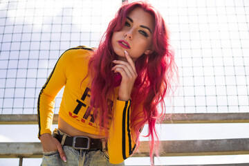 A lady with pink hair in a yellow top, torn jeans and sunglasses stands on the street against the sky. Vintage retro style clothing from the 2000s in modern fashion. The return of old trends.