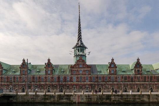 Outdoor exterior front view of B&oslash;rsen, waterfront building  former stock exchange with a striking spire, and people cycling on bicycle lane beside canal in Copenhagen, Denmark. 

