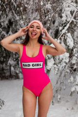 A joyful young lady in a pink one-piece swimsuit and a hat stands in a snowy winter forest and shows her tongue. Cheerful winter mood