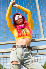 A lady with pink hair, headphones around her neck, in a yellow top, ripped jeans and sunglasses is...