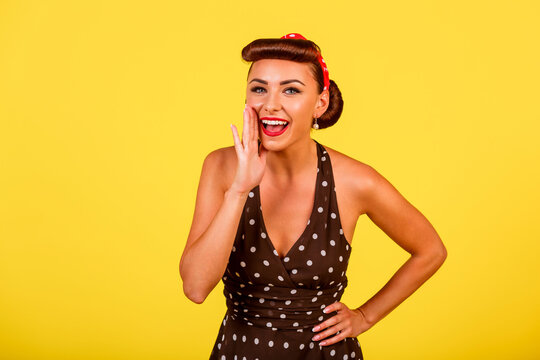 A cute lady in a polka dot dress with a retro hairstyle and red lipstick says something on a yellow background. A pin-up look with a modern twist. Vintage party