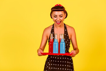 Smiling lady in polka dot dress and retro hairstyle holds a tray with bottles of soda in her hands on a yellow background. A pin-up look with a modern twist. Vintage party