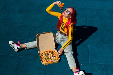 A lady with pink hair in sunglasses, a yellow top and ripped jeans is sitting on the sidewalk eating pizza. Vintage retro clothing style of the 2000s in modern fashion.