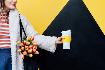 Cropped woman holding reusable coffee cup and fresh tulips in net bag on the geometry bright yellow...